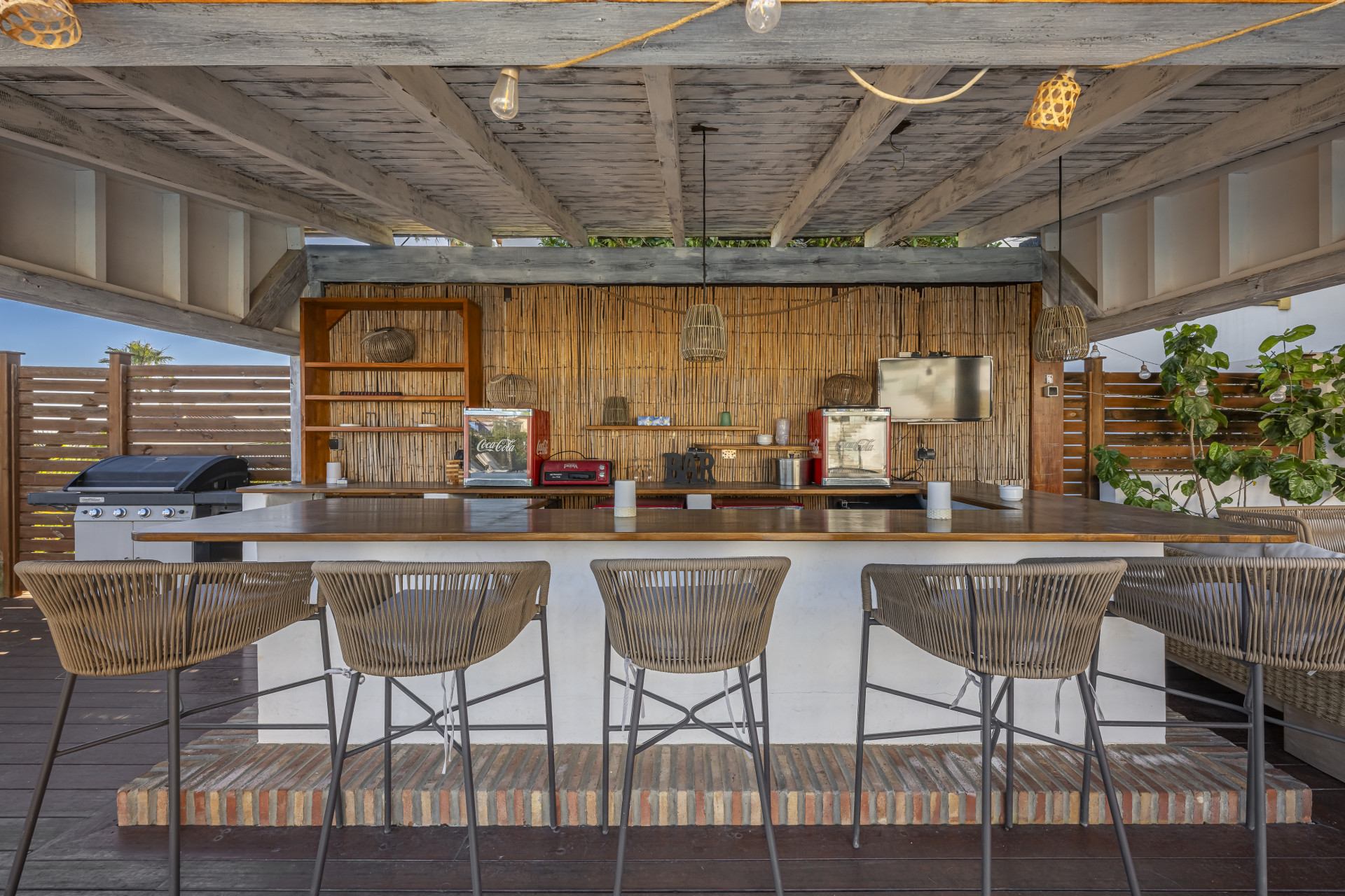 Covered outdoor bar with timber beams, counter seating and a barbecue beside the villa garden.