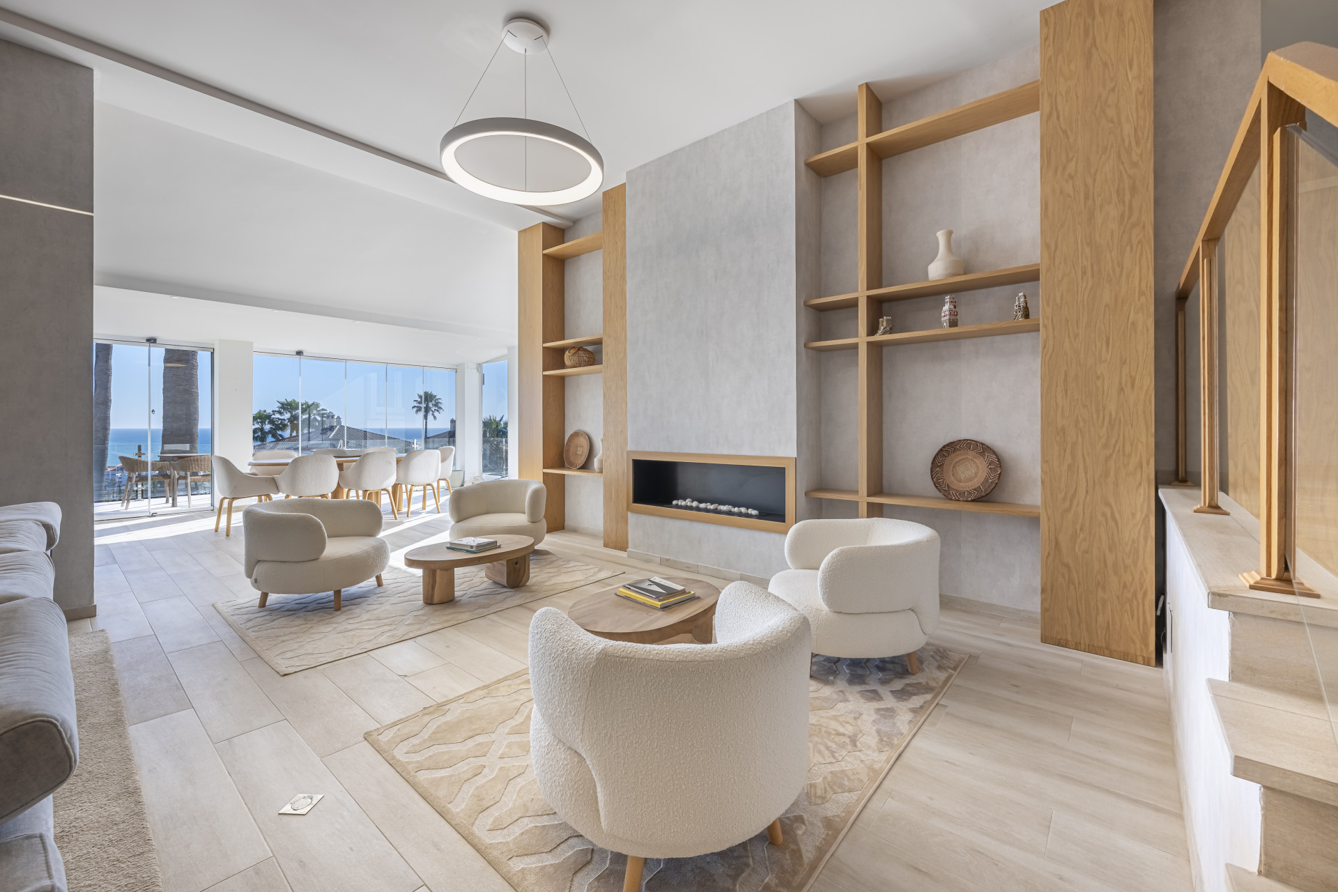 Pale seating and wood shelving around a fireplace, with a glazed dining area and sea views beyond.