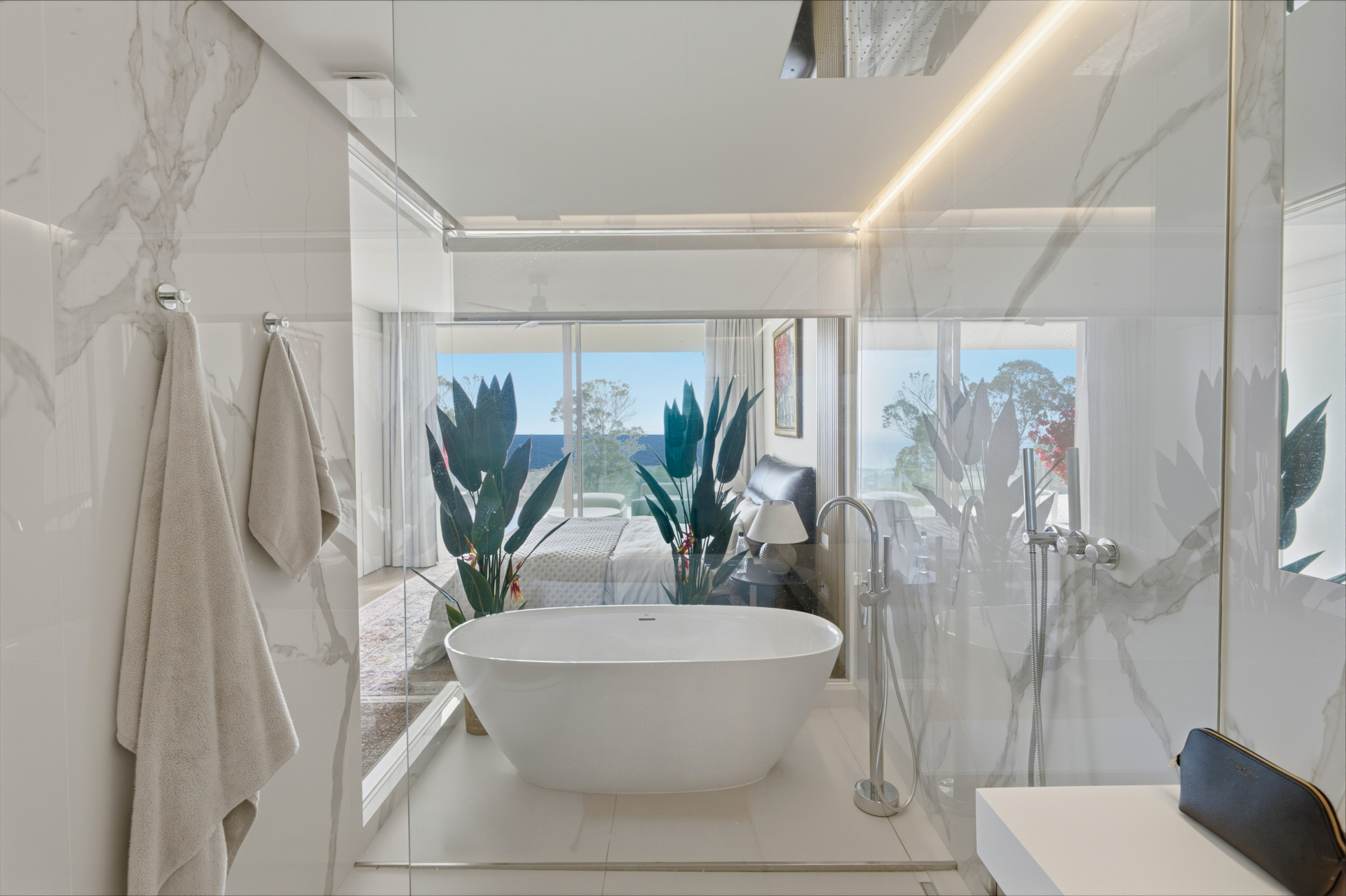 Freestanding bath and glass shower beside a glazed partition looking through the bedroom towards the sea.