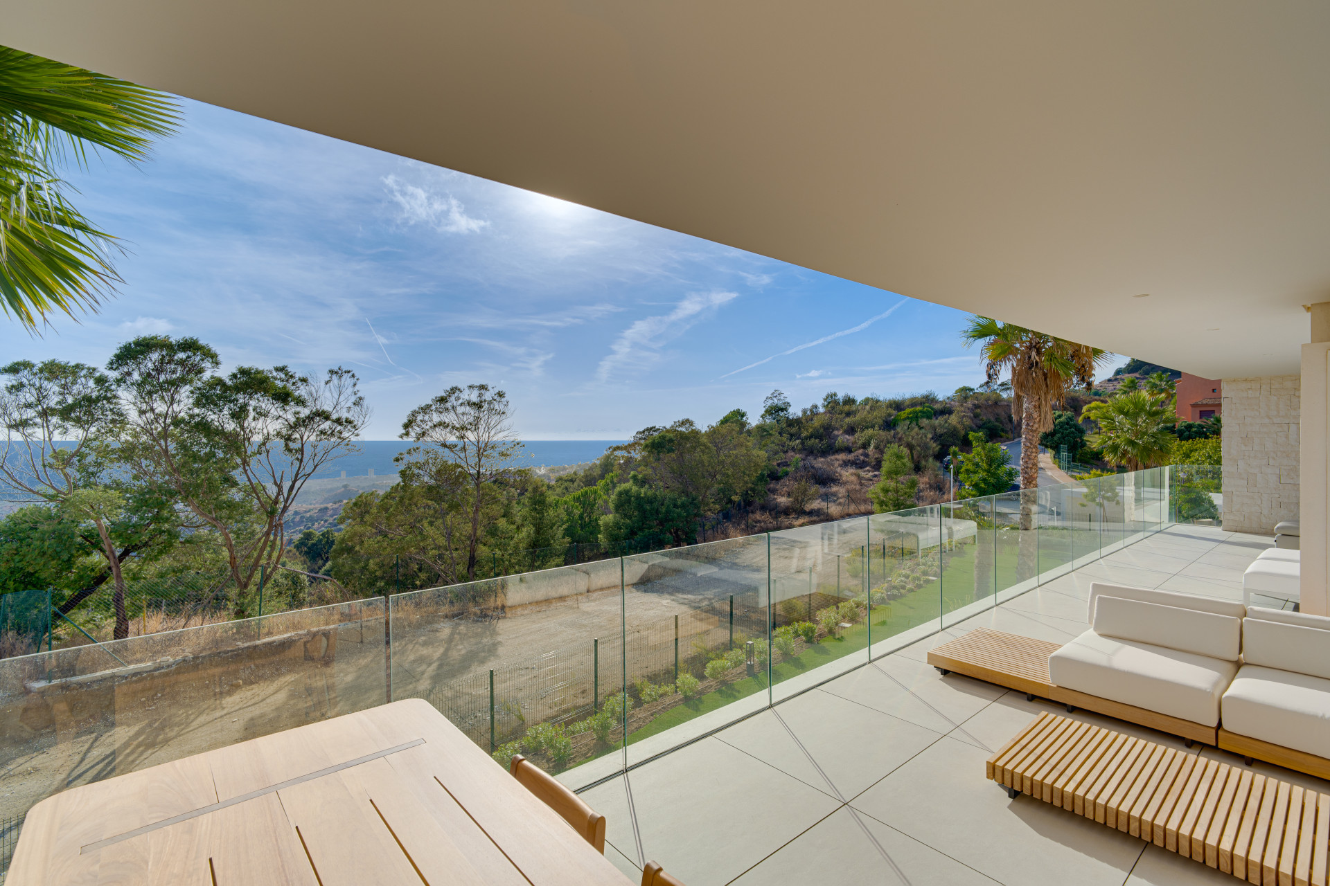 Covered terrace with outdoor seating, a glass balustrade and views across trees to the sea.